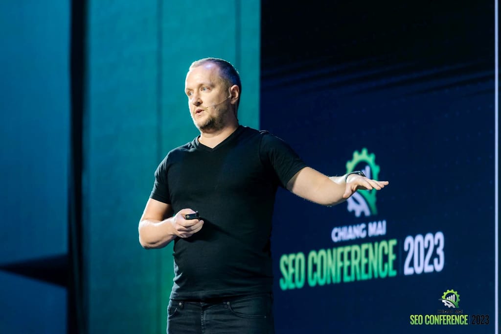 Robert Niechciał gestykuluje podczas wystąpienia na tle logo Chiang Mai SEO Conference 2023