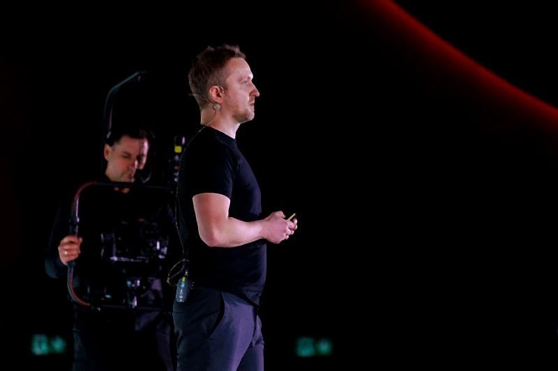 Robert Niechciał z profilu, z mikrofonem nagłownym, na ciemnej scenie I ❤️ AI 2026, za nim operator kamery