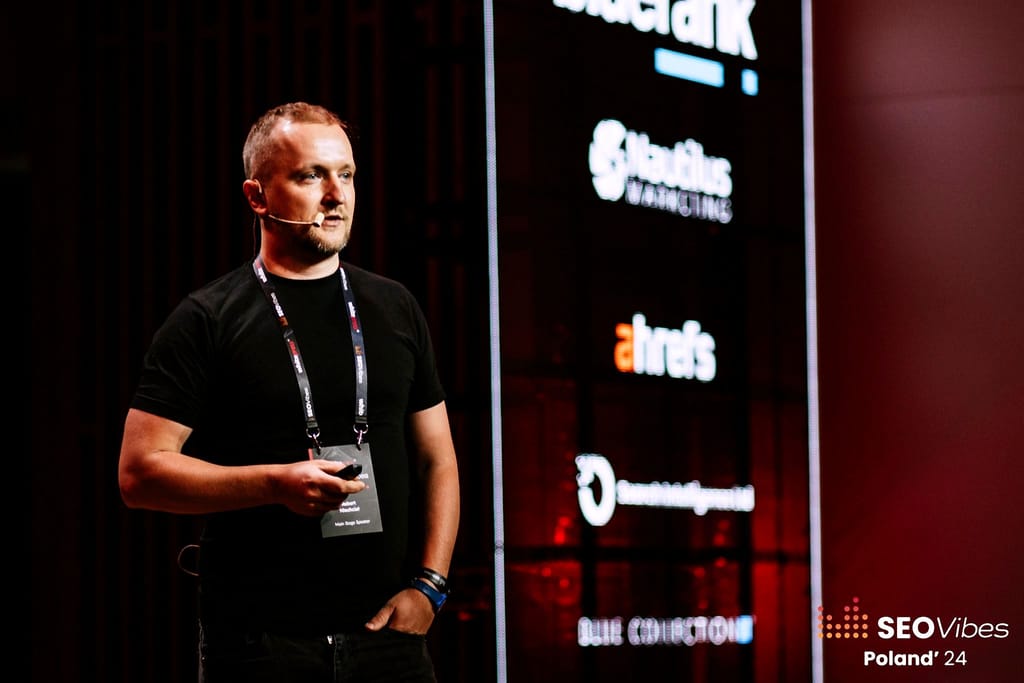 Robert Niechciał z mikrofonem nagłownym na scenie głównej SEO Vibes Poland 2024, za nim ściana sponsorów